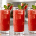 Fresh homemade watermelon juice in a clear glass with ice cubes and a mint garnish on a white marble countertop.