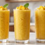 Fresh homemade mango smoothie in a clear glass with a creamy texture, topped with mango cubes and mint on a white marble countertop.