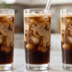 Homemade iced coffee in a clear glass with ice cubes, creamy milk swirl, and a glass straw on a white marble countertop.