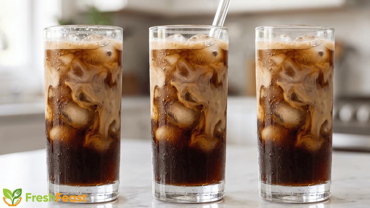 Homemade iced coffee in a clear glass with ice cubes, creamy milk swirl, and a glass straw on a white marble countertop.