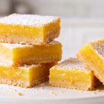 A stack of four homemade lemon bars and two cut pieces dusted with powdered sugar on a white ceramic plate, featuring the FreshFeast logo in the corner.